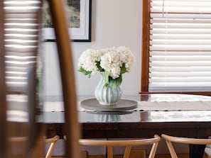 A dining table with a vase of white flowers, a framed picture on the wall, and a window with blinds. - Game Room + Coffee Bar | Near Dining, Parks & Mall (Overland Park)