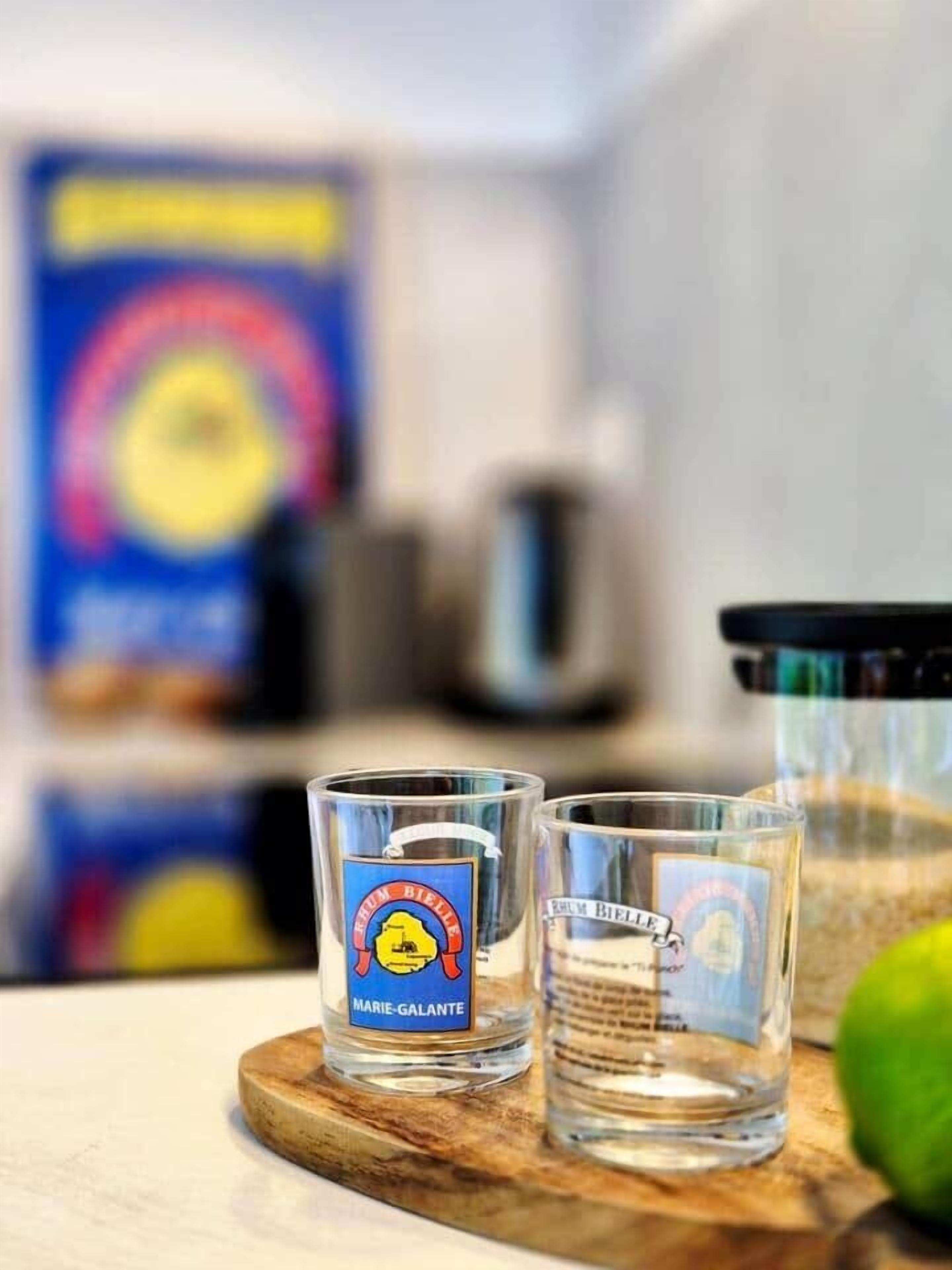Tres copas pequeñas con etiquetas sobre una tabla de madera, una fruta verde y un fondo desenfocado con un objeto azul y amarillo.