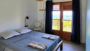 A bedroom with a bed, a wooden bench, a chair, a ceiling fan, and a view through a door. - Villa Bois Coco with a view of Pain de Sucre (Terre-de-Haut)