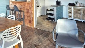 A room with a wooden wall, a dining table with chairs, a kitchen area with cabinets and a refrigerator, and a small cart. - Villa Bois Coco with a view of Pain de Sucre (Terre-de-Haut)