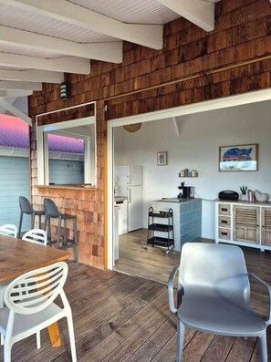 A room with a wooden wall, a dining table with chairs, a kitchen area with cabinets and a refrigerator, and a small cart. - Villa Bois Coco with a view of Pain de Sucre (Terre-de-Haut)