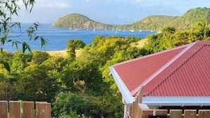 A wooden gate leading to a building with a red roof, surrounded by lush greenery and a view of the sea. - Villa Bois Coco with a view of Pain de Sucre (Terre-de-Haut)