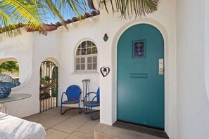 Un edificio bianco con una porta color turchese, due sedie blu, un tavolino e una finestra decorata con una forma a cuore.
