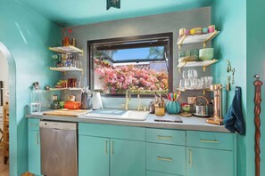 Cucina con mobili color petrolio, lavastoviglie, una finestra vista su fiori rosa e diversi oggetti da cucina sugli scaffali.
