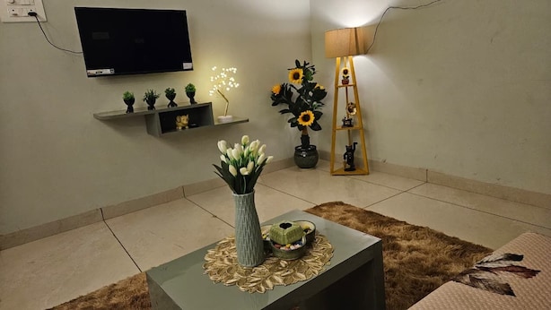 Un salon avec un téléviseur à écran plat, une petite étagère ornée d’objets décoratifs, un vase avec des fleurs, une lampe et un canapé.