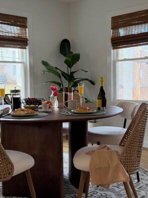 A dining area with a round table, wicker chairs, a potted plant, and breakfast items.