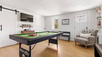 Una mesa de billar con bolas, un televisor, un sofá y una estantería en una habitación con piso de madera y paredes blancas.