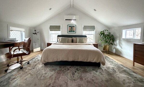 A bedroom with a sloped ceiling, a bed with a patterned bedspread, a desk with a chair, a potted plant, and a window with blinds.
