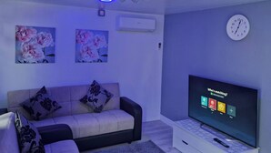 Ein Wohnzimmer mit einer Couch, einem Flachbildfernseher, einer Wanduhr und Deckenleuchten.