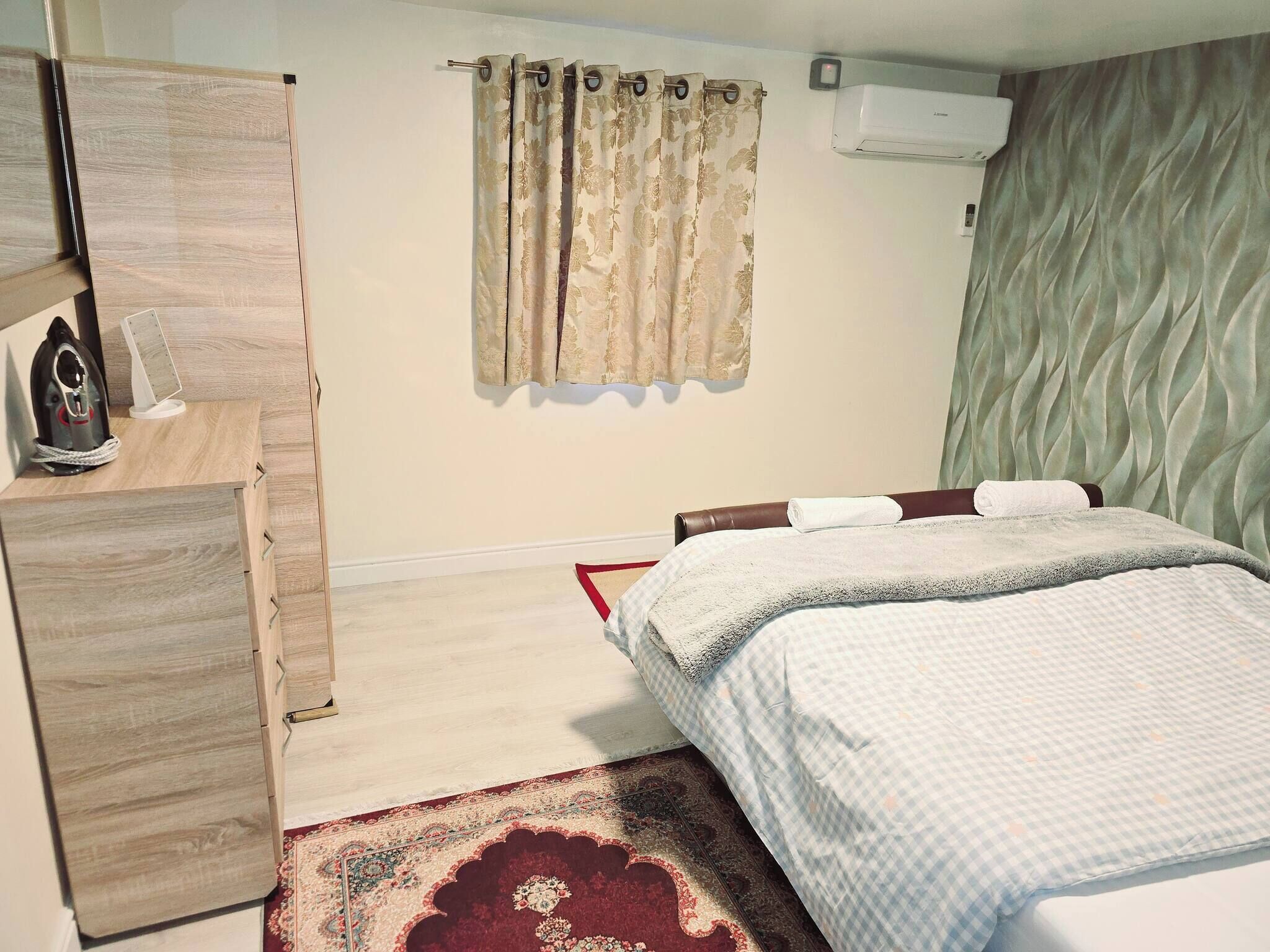 A bedroom with a bed, a wooden dresser, a wall-mounted air conditioner, and a patterned curtain.