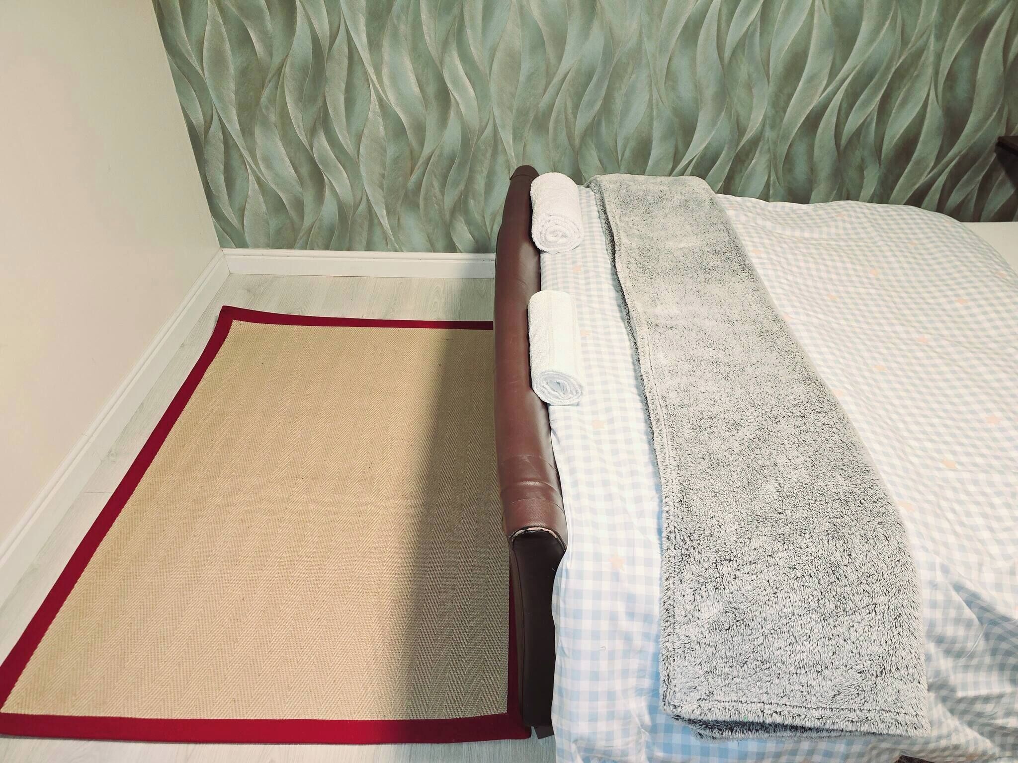 A bed with a checkered bedspread, a rolled-up towel, and a beige rug with a red border.