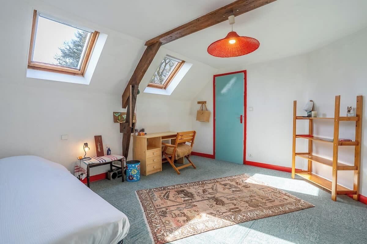 Ein Zimmer mit einem Bett, einem Schreibtisch, einem Sessel, einem Bücherregal, einem Teppich und Dachfenstern.