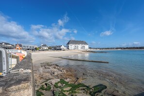 Vista costera con playa arenosa, un muelle con barcos y un edificio con tejado inclinado.