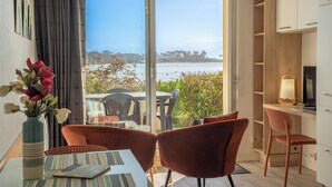 A room with a sliding glass door leading to a balcony with a view of the sea, a dining table with chairs, a TV, and a bookshelf. - Sea-view garden-level flat (Ploemeur)