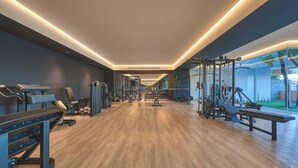 Academia moderna com piso de madeira, diversos equipamentos de exercício e vista para o exterior através de amplas janelas.