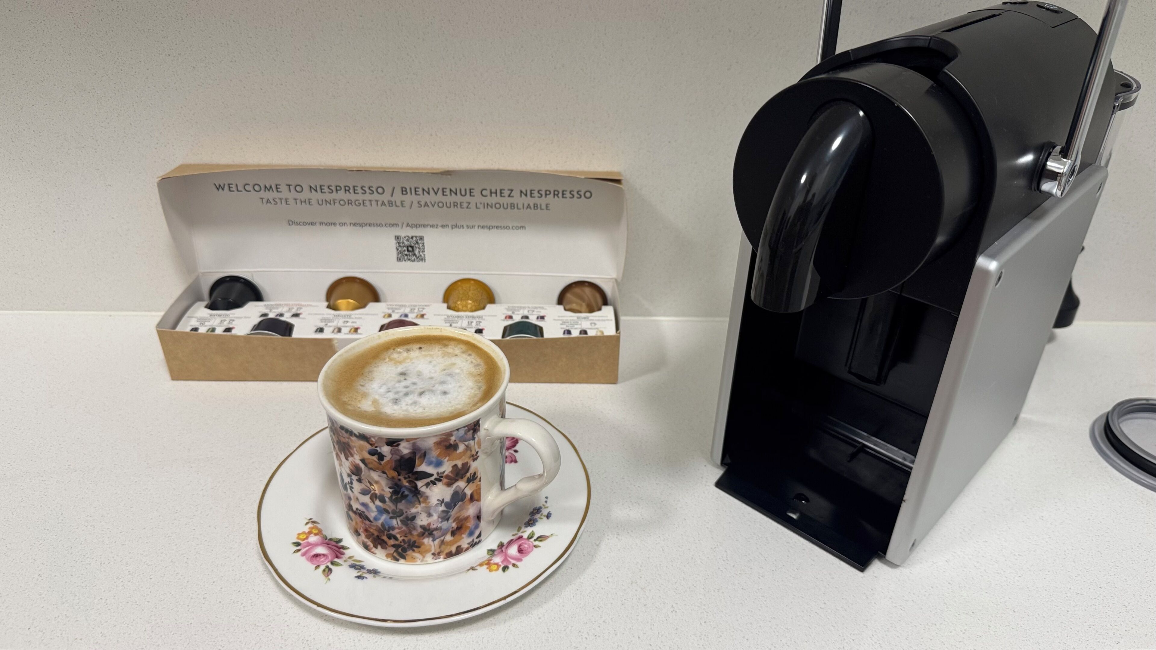 Kahvinkeitin, kuppi kahvia ja paketti Nespresso-kahvikapseleita keittiötasolla.