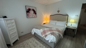 Una camera da letto con un letto, un comodino con una lampada, una cassettiera, un quadro appeso al muro e una decorazione a forma di cuore.