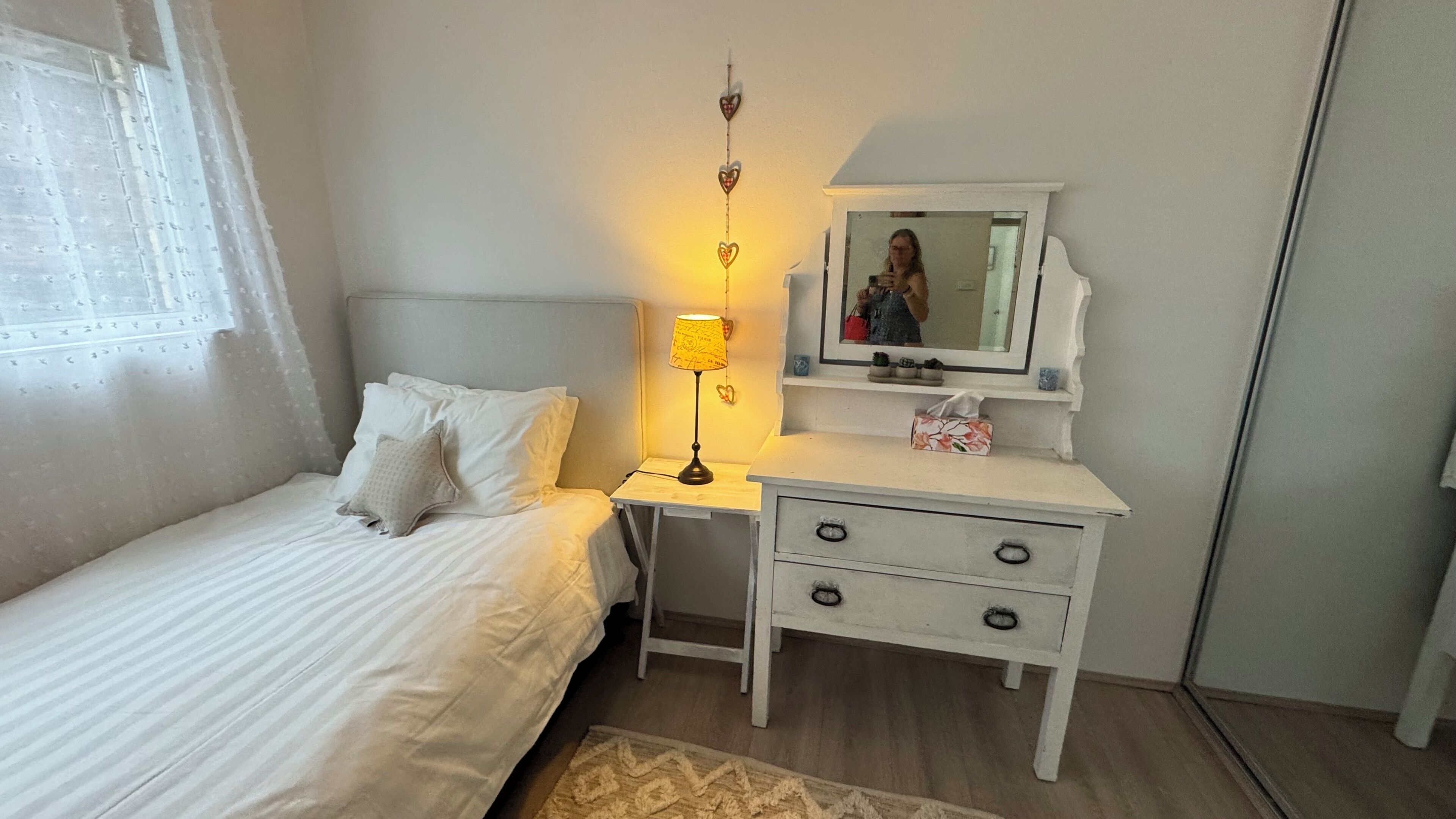 A bedroom with a bed, bedside table with a lamp, a dresser with drawers, a mirror, and a window with curtains.