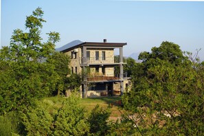 A two-story stone house with a balcony, surrounded by dense greenery and a mountain in the background. - Cozy Apartment with Breathtaking Views, Private Terrace Wi-Fi & Air Conditioner (Petrovac)