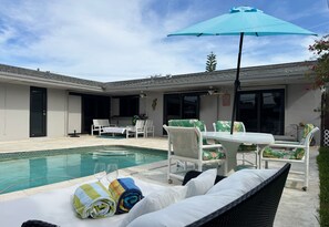 Et poolområde med et bord og stole, en blå parasol og et hvidt udendørs loungeområde.
