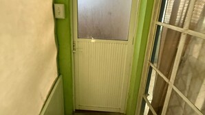 Een smalle gang met een groene muur, een witte deur met een geëtste ruit en een raam met een metalen rooster.