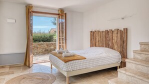 Ein Schlafzimmer mit einem Bett, einem hölzernen Kopfende, einer Steinwand und Blick durch eine offene Tür.