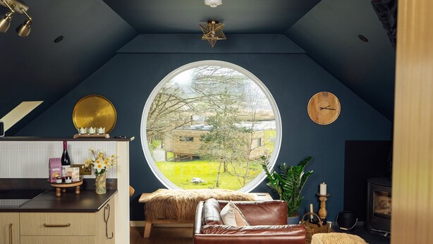 A cozy living room with a large round window, a fireplace, and a kitchen area with a bar. - Riverside living, reimagined above the Teifi (Cardigan)