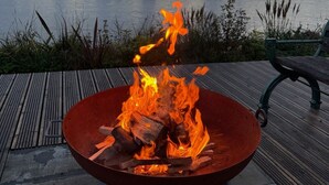 A fire pit with logs, a bench, and a view of a body of water and hills. - Riverside living, reimagined above the Teifi (Cardigan)