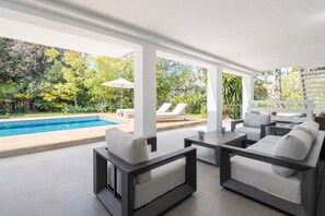 A modern outdoor lounge area with a pool, white furniture, and lush greenery. - Villa Karma -Elegant Villa with Pool, Garden & Mul (Benahavís)
