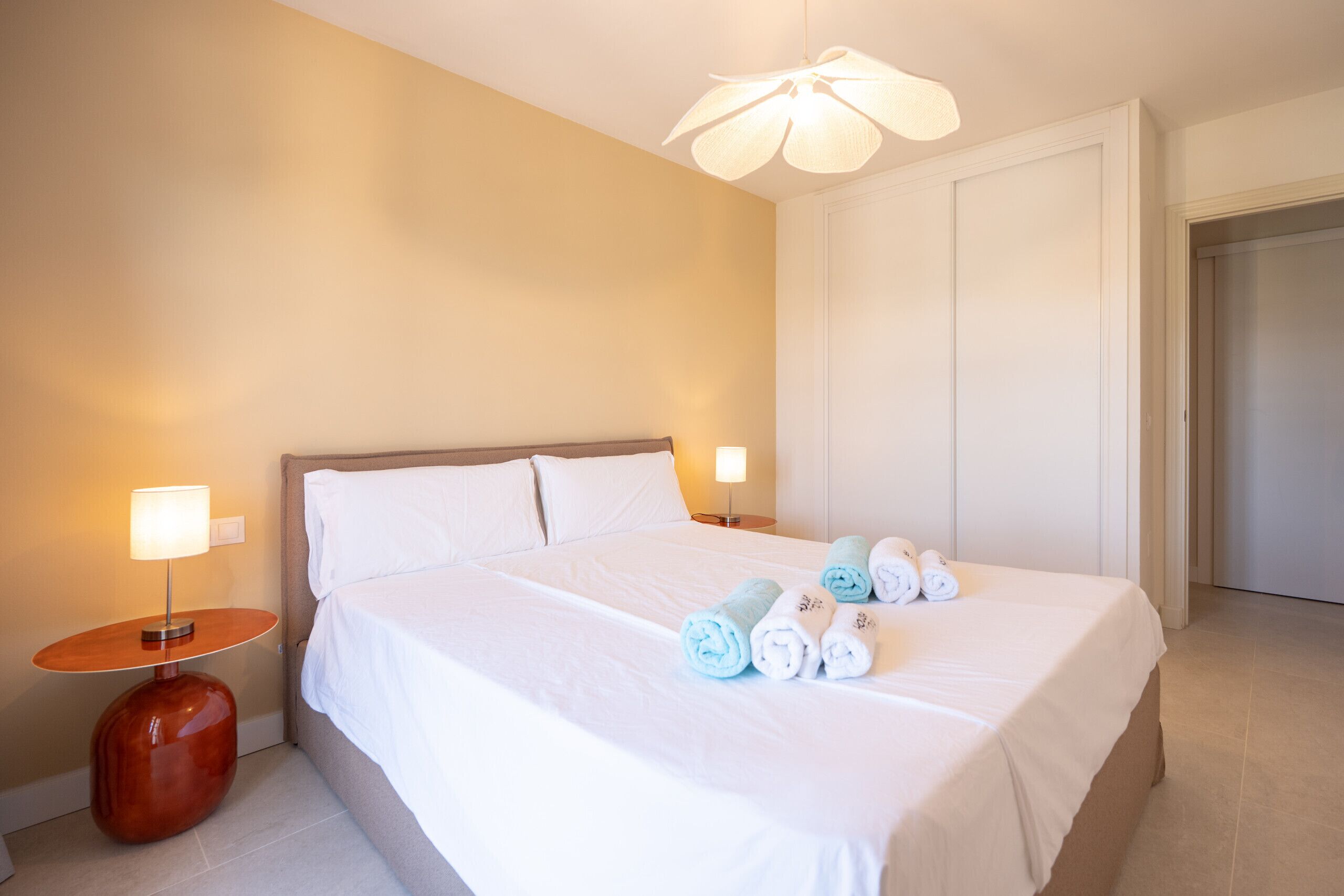 A hotel room with a large bed, two bedside lamps, and neatly rolled towels on the bed.