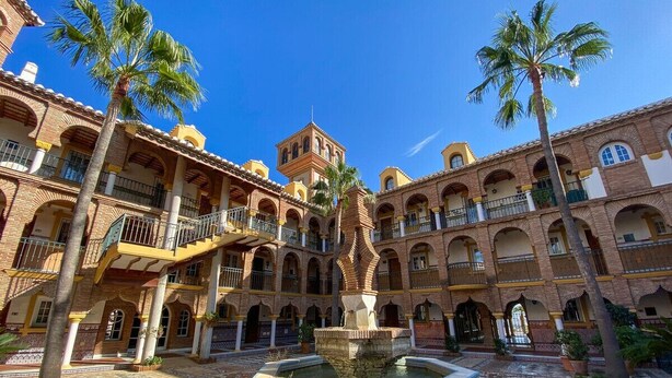 A courtyard with a fountain, palm trees, and a building with multiple balconies and arches. - Puebla Aida Unique - Charming Renovated 2 bedroom (Mijas Costa)