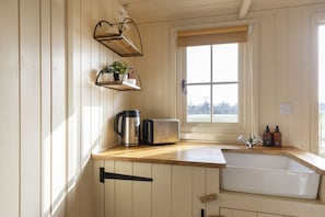 A kitchen with wooden cabinets, a sink, a toaster, a kettle, and a window with a view. - Riverside Shepherd's Hut with Hot Tub | Tewkesbury (Tewkesbury)