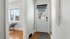 A hallway with a white door, a gray chair, and a view of the outside. - Comfortable Crescentwood Bungalow | 3 Bed | 2 Bath | (Winnipeg)