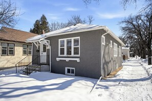 Una casa residenziale con facciata grigia, infissi bianchi e un portico con ringhiera nera, oltre a un vialetto ricoperto di neve.