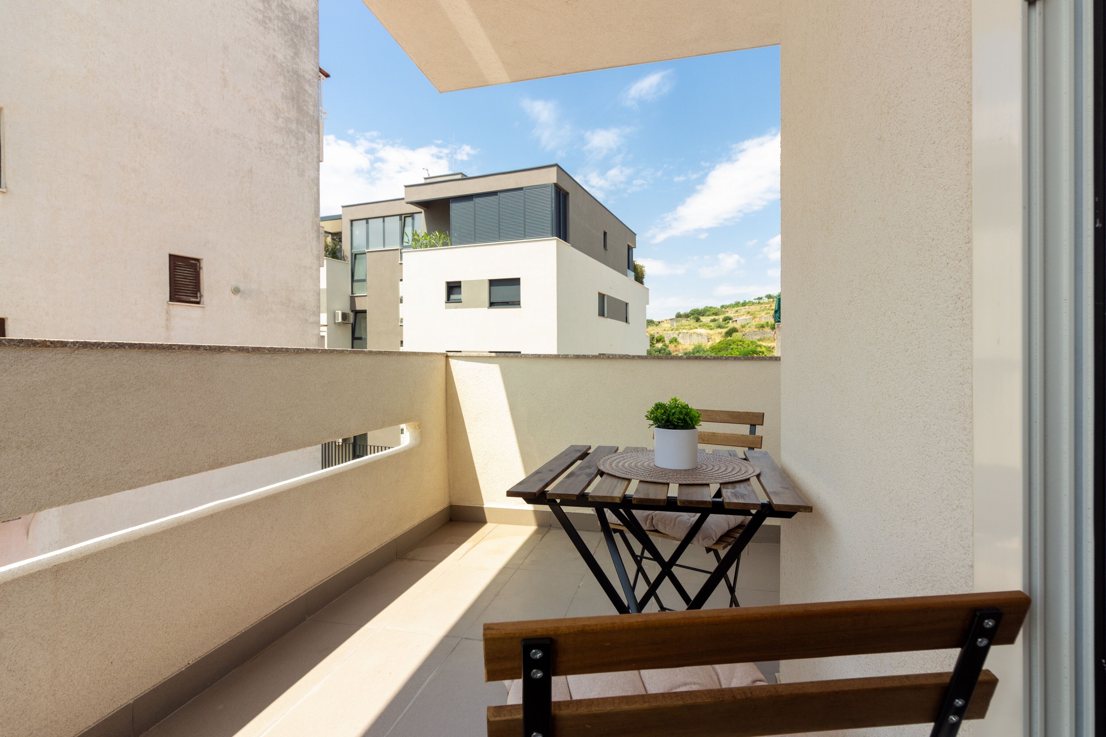 Un balcon avec un banc en bois, une petite table sur laquelle est posée une plante en pot, et une vue sur des immeubles modernes.