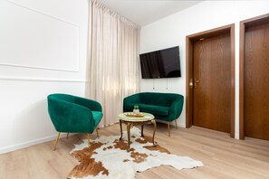 A room with two green armchairs, a small round table with a plant, a mounted TV, and a wooden door.
