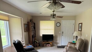 Ein Wohnzimmer mit Deckenventilator, Fernseher, Bücherregal, einer Tür und einem Fenster mit Jalousien.