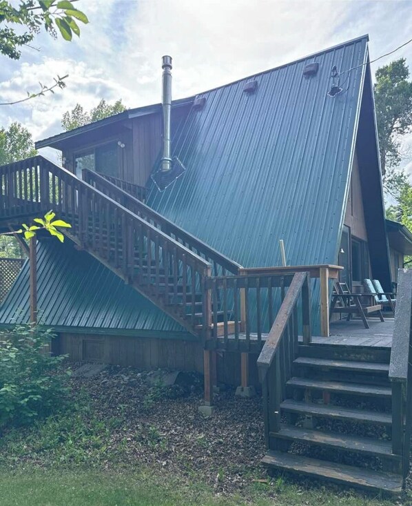A rustic cabin with a steeply pitched metal roof, a wooden staircase, and a deck with outdoor seating.