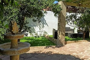 A stone patio with a fountain, a white wall, and a seating area with chairs.