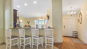 Een keuken met een bar, witte stoelen, een kroonluchter en een deur naar een andere kamer.