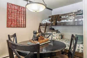 Une chambre confortable, aménagée dans le style d’un chalet, avec une table de salle à manger ronde, des chaises en bois, un fauteuil suspendu, une figurine d’ours et une pancarte de bienvenue.