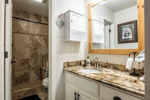 Une salle de bain dotée d’un comptoir en granit, d’un lavabo blanc, d’un miroir avec un cadre en bois et d’un espace douche aux carreaux beiges.