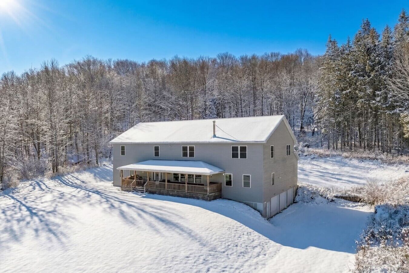 Une maison à deux étages, dotée d’un porche couvert et d’un garage, entourée d’un paysage enneigé et d’une forêt dense.