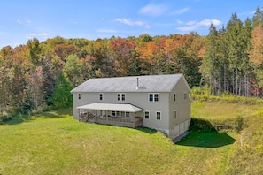 A two-story house with a covered porch, surrounded by a grassy yard and dense trees. - Spacious 9BR Catskills Retreat • Views + Game Room (Andes)