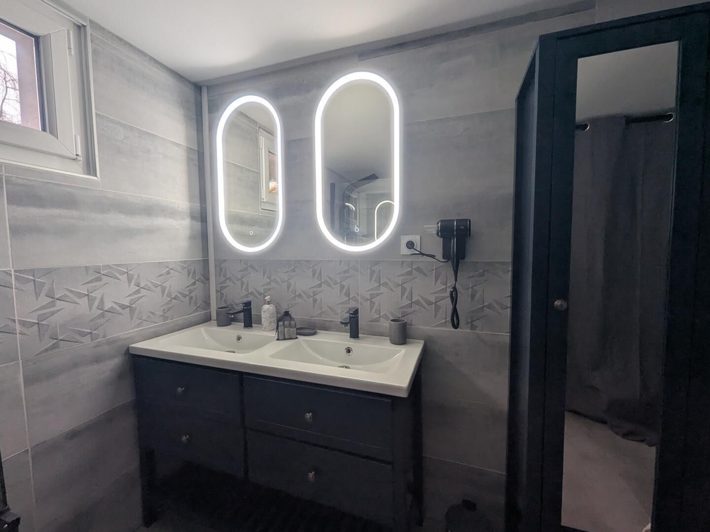Ein Badezimmer mit zwei Waschbecken, einem Waschtisch mit Schubladen, einem Spiegel mit beleuchtetem Rand und einem an der Wand montierten Föhn.