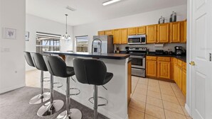 Eine moderne Küche mit Holzschränken, einem Edelstahl-Kühlschrank, einer Mikrowelle und einem eingebauten Backofen. An einer zentralen Kochinsel stehen drei schwarze Barhocker.