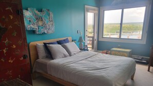 A bedroom with a bed, a dresser, a window with a view, and artwork on the wall.