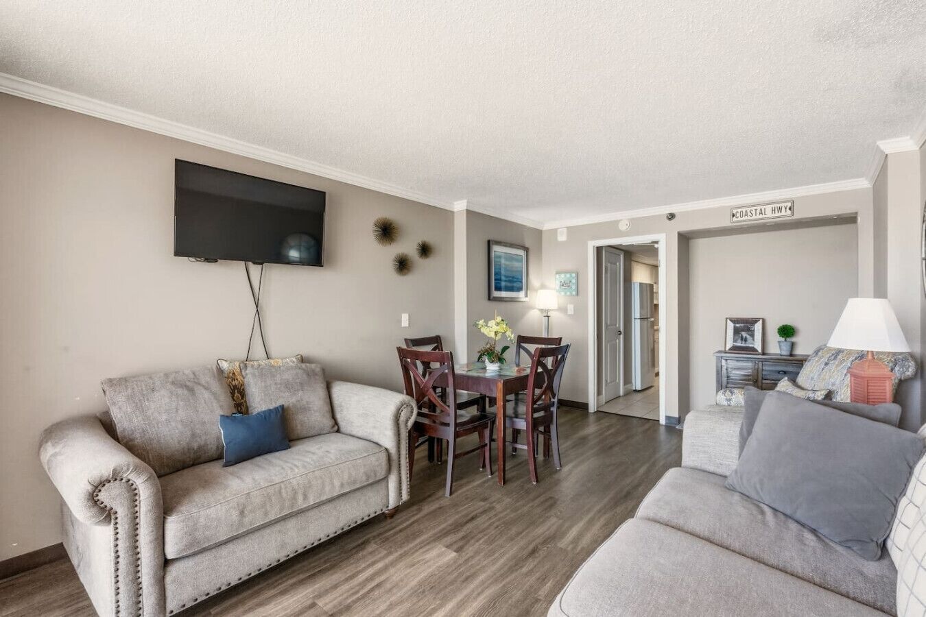 A living room with a sofa, a dining table, and chairs. There is a flat-screen TV mounted on the wall.