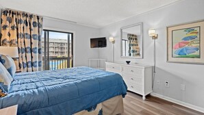A bedroom with a bed, a dresser, a TV, a window with curtains, and a painting on the wall. - Beachfront Resort Condo with Pools and Hot tub (North Myrtle Beach)