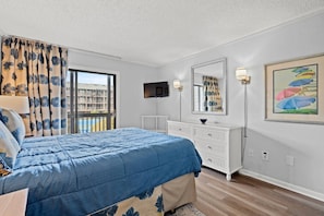 Un dormitorio con una cama, un aparador, un televisor, una ventana con cortinas y un cuadro en la pared.
